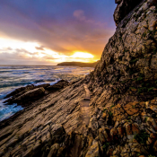 Rocky coastline of Robberg Nature Reserve with rugged cliffs, waves crashing against the shore, and a sandy peninsula stretching into the ocean under a gorgeous sky.