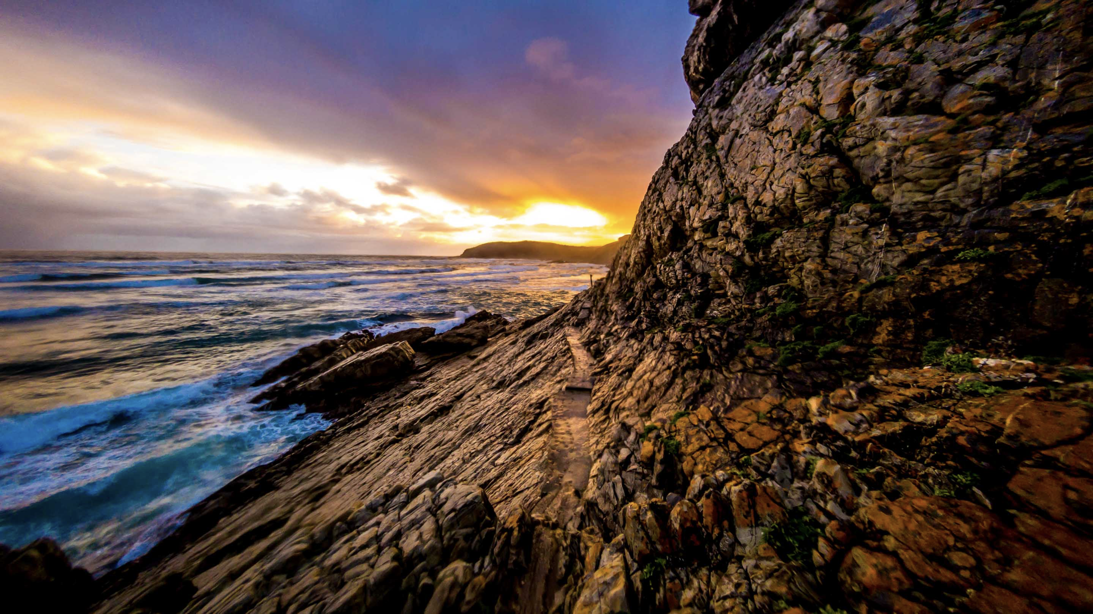 Rocky coastline of Robberg Nature Reserve with rugged cliffs, waves crashing against the shore, and a sandy peninsula stretching into the ocean under a gorgeous sky.